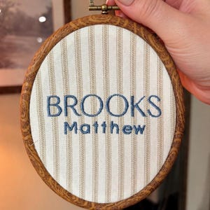 May include: A circular embroidery hoop with a faux wood grain frame containing cream-colored fabric with vertical tan stripes. The name BROOKS Matthew is embroidered in the center in a dark blue thread. A small metal hanging loop is at the top.