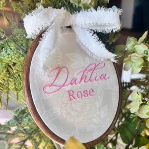 May include: An oval wooden embroidery hoop frame containing off-white fabric with a subtle floral pattern. The name Dahlia Rose is embroidered in pink thread in the center. A frayed white ribbon is tied in a bow at the top of the hoop.
