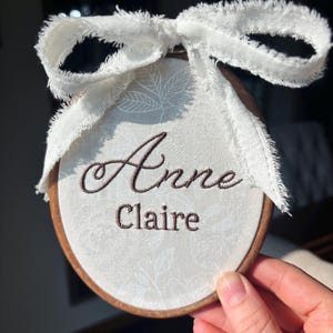 May include: A circular wooden embroidery hoop containing white fabric with a subtle floral pattern. The name Anne Claire is embroidered in dark brown thread. A white frayed ribbon is tied in a bow at the top of the wooden frame.