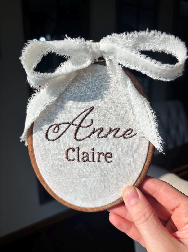 May include: A circular wooden embroidery hoop containing white fabric with a subtle floral pattern. The name Anne Claire is embroidered in dark brown thread. A white frayed ribbon is tied in a bow at the top of the wooden frame.