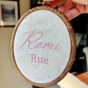 May include: An oval wooden embroidery hoop frame containing light beige fabric with a white floral pattern. The names Remi and Rue are embroidered in pink thread in the center of the fabric. The hoop features a metal tightening screw.