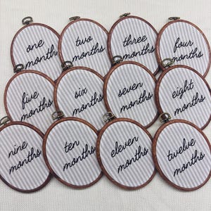 May include: A set of twelve oval embroidery hoops featuring white and gray vertical striped fabric. Each hoop contains black embroidered text ranging from one month to twelve months, with a small metal tightening screw at the top.