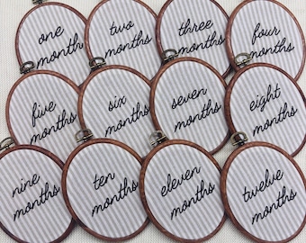 May include: A set of twelve oval embroidery hoops featuring white and gray vertical striped fabric. Each hoop contains black embroidered text ranging from one month to twelve months, with a small metal tightening screw at the top.