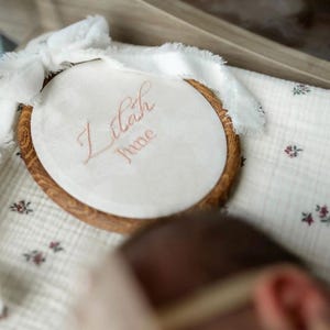 May include: An oval wooden embroidery hoop containing white fabric with the embroidered name Lilah June in light pink script. A white frayed ribbon is tied in a bow at the top of the hoop, which rests on a white fabric with a small floral pattern.