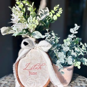 May include: An oval wooden embroidery hoop containing cream fabric with white floral lace patterns, embroidered with the name Lilah and the month June in red thread, accented by a frayed white ribbon bow and artificial green foliage.