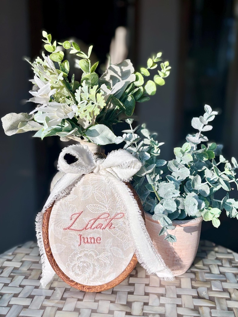 May include: An oval wooden embroidery hoop containing cream fabric with white floral lace patterns, embroidered with the name Lilah and the month June in red thread, accented by a frayed white ribbon bow and artificial green foliage.