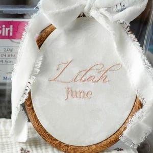 May include: A circular wooden embroidery hoop containing white fabric with the embroidered names Lilah and June in light pink thread. A white frayed ribbon is tied in a bow at the top of the hoop. The fabric features a subtle floral pattern.