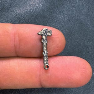 May include: A small, bronze-colored pendant in the shape of a hammer. The hammer head has a textured design, and the handle has a branch-like appearance. A small loop is at the bottom of the handle.