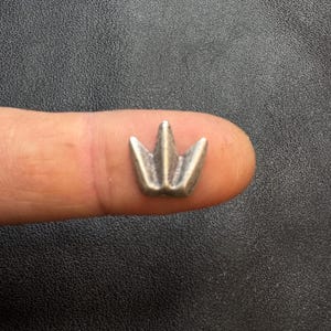May include: A small, silver-colored crown charm with three points. The charm is held on a finger against a dark background. The crown is a miniature decorative element, likely for jewelry or crafts.