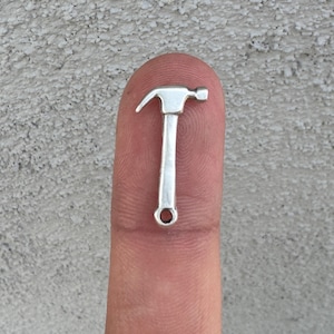 Puede incluir: Un pequeño charm de martillo de plata con una cabeza redonda y un agujero en la parte inferior para sujetarlo a una cadena o pulsera.