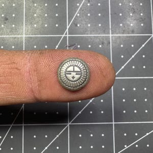 Op de afbeelding: Een kleine, ronde, zilverkleurige knoop met een zonachtig ontwerp in het midden. De knoop heeft een gedetailleerd, reliëfpatroon rond de rand en wordt op een vingertop tegen een donkere achtergrond gehouden.