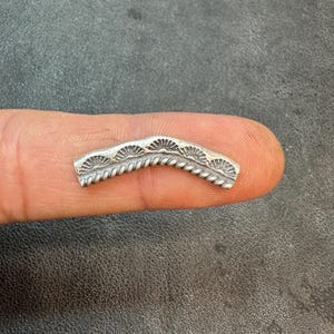 May include: A small, curved silver metal piece with a decorative design. The piece features a series of fan-like shapes along the top edge and a rope-like pattern along the bottom edge. The item is held on a finger against a dark background.