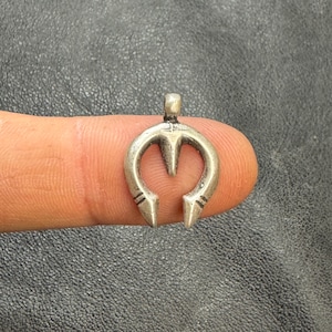 May include: A small, silver-colored pendant with a unique horseshoe-like design. The pendant features a central, pointed element and two outward-facing, pointed ends. The piece is held up by a small loop at the top, suitable for a chain or cord.