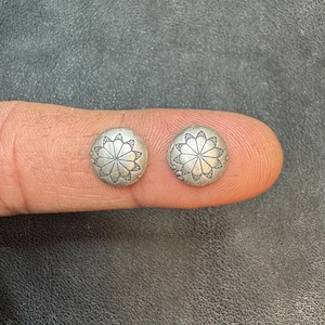 Op de afbeelding: Twee kleine, ronde zilverkleurige oorstekers. Elke oorbel heeft een gedetailleerd bloemmotief in het midden. De oorbellen worden op een vinger getoond, waarbij hun grootte en ontwerp worden benadrukt.