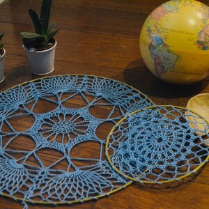 May include: Two blue crochet doilies with a circular design, one large and one small, are displayed on a wooden surface. A globe, two small potted succulents, and a decorative leaf-shaped object are also present.
