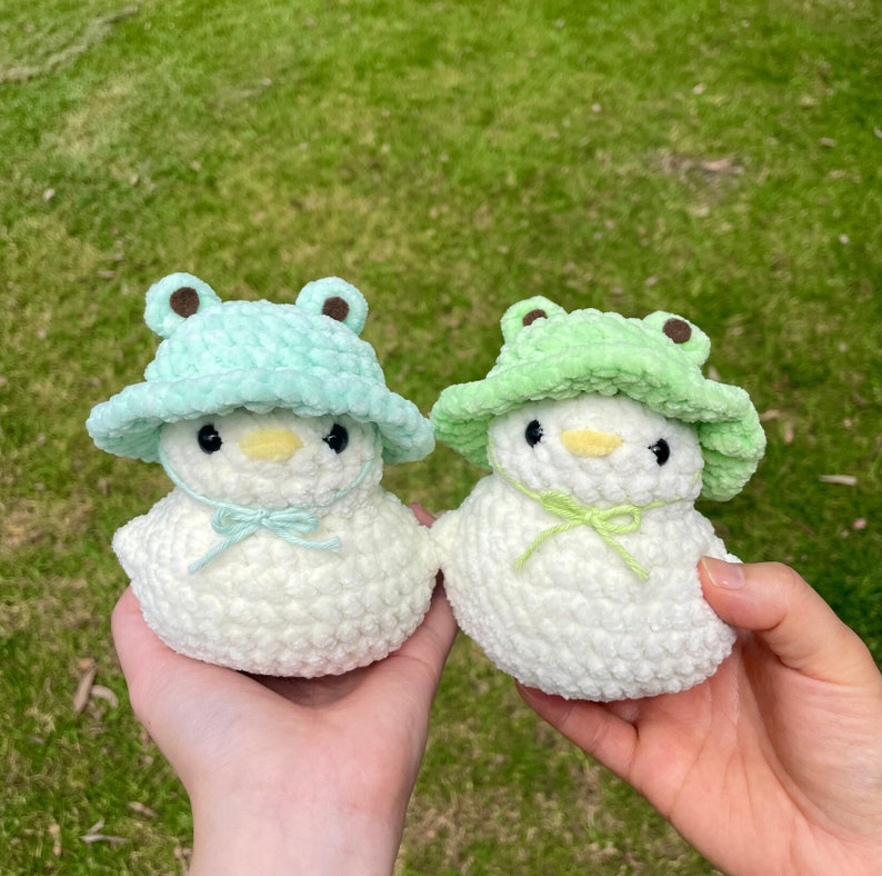 May include: Two white crocheted plush toys shaped like chicks wearing green crocheted frog hats. The hats have brown felt eyes and a wide brim.
