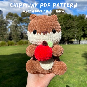 May include: A brown and white crocheted chipmunk plush toy holding a red apple. The toy has black eyes and a white nose. The background is a blurry image of a park.