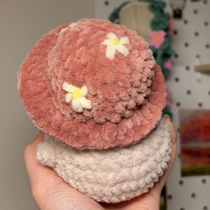 K&ouml;nnte beinhalten: Ein rosa geh&auml;kelter Bucket-Hut mit wei&szlig;en G&auml;nsebl&uuml;mchen-Verzierungen sitzt auf einer kleineren, hellrosa geh&auml;kelten Kugel.