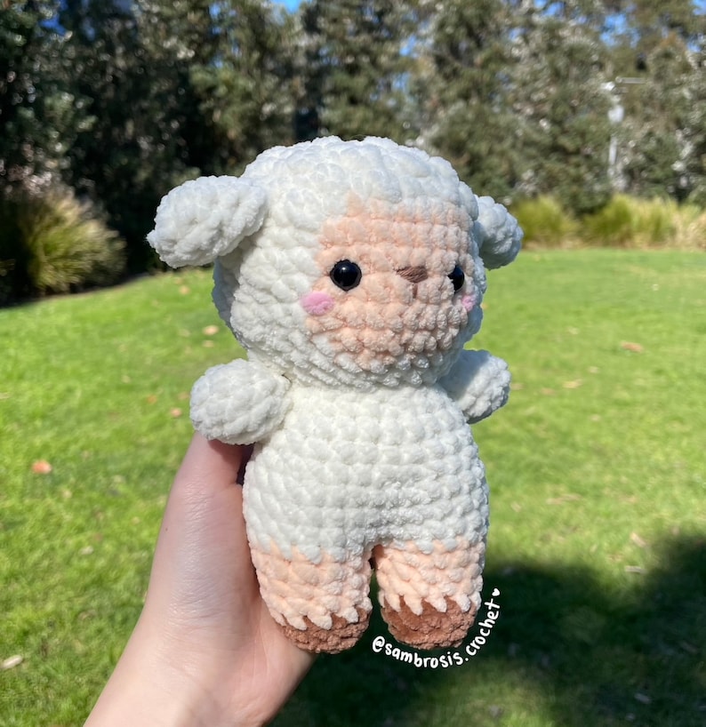 May include: A white crocheted lamb plush toy with brown hooves and a pink nose. The lamb has black eyes and pink cheeks. The toy is being held in a hand.