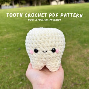 May include: A crocheted tooth plushie in cream-colored yarn, featuring black eyes, a small smile, and pink cheeks. The text "TOOTH CROCHET PDF PATTERN" and "*NOT A PHYSICAL PLUSHIE*" are displayed above the plushie.