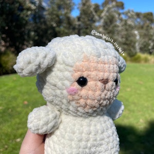 May include: A white crocheted lamb with a pink nose and black eyes. The lamb is holding its head up and looking to the right. The lamb is sitting on a green grassy surface. The text "@sambrosis.crochet" is visible in the upper right corner of the image.
