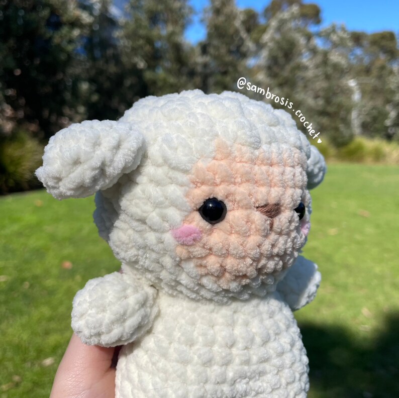 May include: A white crocheted lamb with a pink nose and black eyes. The lamb is holding its head up and looking to the right. The lamb is sitting on a green grassy surface. The text "@sambrosis.crochet" is visible in the upper right corner of the image.