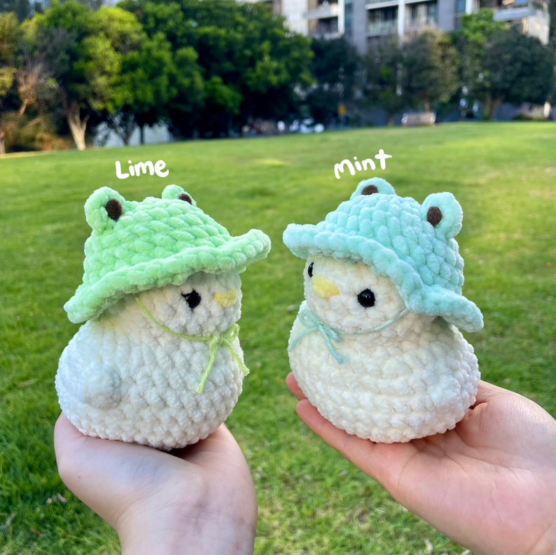 May include: Two white crocheted plush toys wearing green frog hats. The hat on the left is lime green and the hat on the right is mint green. The text "Lime" is above the left toy and the text "Mint" is above the right toy.