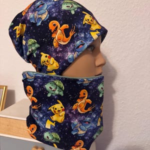 Könnte beinhalten: Ein Set aus einer dunkelblauen Mütze und einem passenden Schlauchschal mit einem wiederkehrenden Muster aus bunten Zeichentrickfiguren, darunter gelbes Pikachu, orangefarbenes Glumanda, blaues Schiggy und grünes Bisasam auf sternenhimmelartigem Grund.