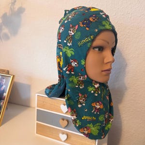 Könnte beinhalten: Eine türkisfarbene Stoffkapuze mit einem sich wiederholenden Muster aus Paw Patrol-Figuren in Dschungel-Outfits, grünen tropischen Blättern und dem Schriftzug Jungle Pups. Die Kapuze bedeckt Kopf und Hals und hat eine Öffnung für das Gesicht.