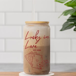 May include: A glass beverage container with a wooden lid and a clear straw. The container has the words "Lucky in Love" and "EST. 2026" in red script. Below the text are red illustrations of playing cards and dice.