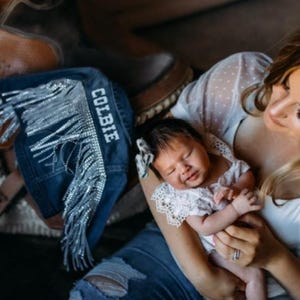 Puede incluir: Una mujer con un vestido blanco de lunares sostiene a un beb&eacute; que lleva un vestido blanco de encaje. El beb&eacute; est&aacute; durmiendo y la mujer est&aacute; mirando al beb&eacute;. La mujer est&aacute; sentada en una silla de montar de cuero marr&oacute;n con una franja de mezclilla azul con la palabra "COLBIE" bordada.