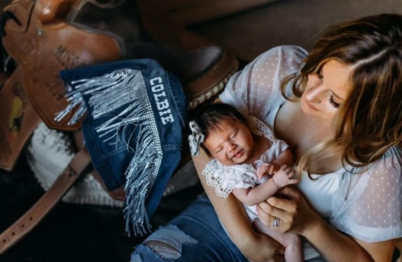 Puede incluir: Una mujer con un vestido blanco de lunares sostiene a un beb&eacute; que lleva un vestido blanco de encaje. El beb&eacute; est&aacute; durmiendo y la mujer est&aacute; mirando al beb&eacute;. La mujer est&aacute; sentada en una silla de montar de cuero marr&oacute;n con una franja de mezclilla azul con la palabra "COLBIE" bordada.