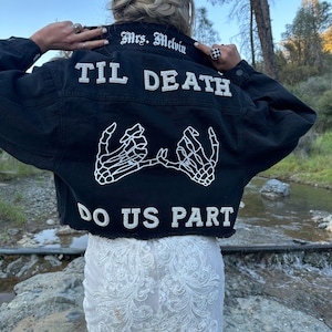 Puede incluir: Una chaqueta vaquera negra con bordado blanco que dice "Mrs. Melvin Til Death Do Us Part" y presenta un par de manos de esqueleto que se toman de la mano con anillos en los dedos.