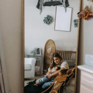 Puede incluir: Una mujer est&aacute; sentada en una alfombra frente a un espejo, sosteniendo un beb&eacute;. El espejo refleja una habitaci&oacute;n de beb&eacute; con una cuna, una mecedora y una chaqueta vaquera con el nombre "Colbie" en la espalda.