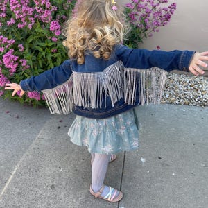 Puede incluir: Una persona joven lleva una chaqueta vaquera azul con flecos plateados y una falda floral azul claro. La chaqueta tiene un ribete brillante.