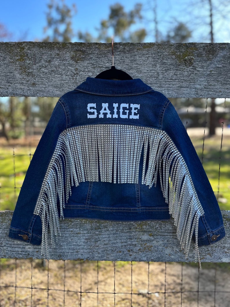 Puede incluir: Una chaqueta vaquera azul con flecos plateados y el nombre "Saige" bordado en la espalda.