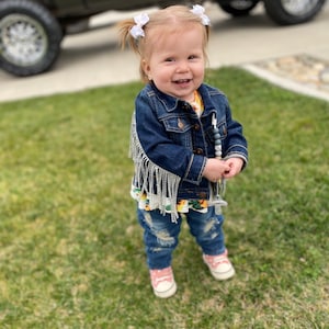 Puede incluir: Una ni&ntilde;a peque&ntilde;a con una chaqueta vaquera azul con flecos plateados, pantalones vaqueros rotos y zapatillas rosas. Est&aacute; sonriendo y lleva lazos blancos en el pelo.