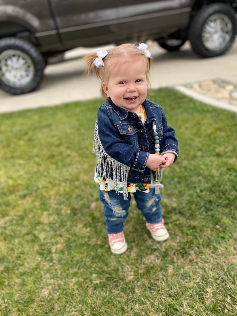 Puede incluir: Una ni&ntilde;a peque&ntilde;a con una chaqueta vaquera azul con flecos plateados, pantalones vaqueros rotos y zapatillas rosas. Est&aacute; sonriendo y lleva lazos blancos en el pelo.