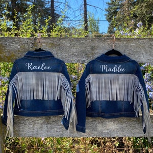 Puede incluir: Dos chaquetas vaqueras con pedrer&iacute;a y flecos. Las chaquetas est&aacute;n colgadas en una valla de madera. Las chaquetas est&aacute;n personalizadas con los nombres "Raelee" y "Madalee".