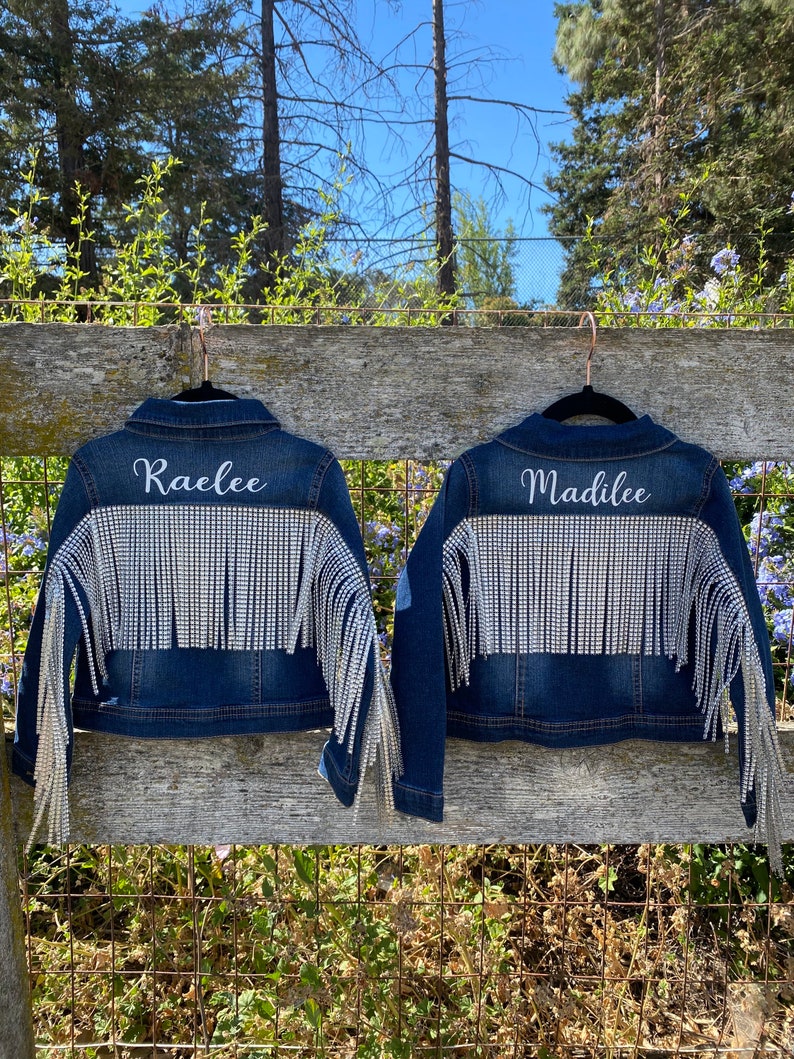 Puede incluir: Dos chaquetas vaqueras con pedrer&iacute;a y flecos. Las chaquetas est&aacute;n colgadas en una valla de madera. Las chaquetas est&aacute;n personalizadas con los nombres "Raelee" y "Madalee".