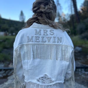 Puede incluir: Chaqueta vaquera blanca con el texto "MRS MELVIN" en pedrería plateada en la espalda. La chaqueta presenta flecos plateados en la espalda y en las mangas. La chaqueta tiene un aspecto desgastado con una sección rasgada en la parte inferior.