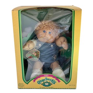 May include: A Cabbage Patch Kids doll from 1985 with blonde yarn hair and blue eyes, wearing blue overalls with an elephant graphic. The doll is inside a yellow cardboard box with a clear plastic window and the Cabbage Patch Kids logo printed.