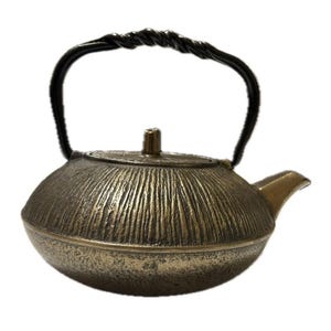 Puede incluir: Una tetera de hierro fundido de color bronce con una superficie texturizada y un asa negra. El asa es arqueada y tiene un diseño retorcido. El pico es pequeño e inclinado. La tetera tiene una tapa con un pequeño pomo.