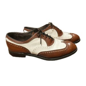 May include: A pair of two-tone wingtip oxford shoes featuring smooth brown leather panels and textured cream leather inserts. The shoes include dark brown laces, decorative brogue perforations along the seams, and a low stacked heel.