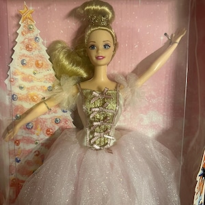 Puede incluir: Muñeca Barbie con un vestido rosa y dorado con una falda de tul blanca. La muñeca tiene el pelo rubio y está en una caja con un diseño de árbol de Navidad. El logotipo de Barbie es visible en la parte inferior de la caja.