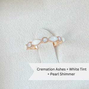 May include: A delicate rose gold ring with white opal stones set in a curved band. The ring is described as "Cremation Ashes + White Tint + Pearl Shimmer".