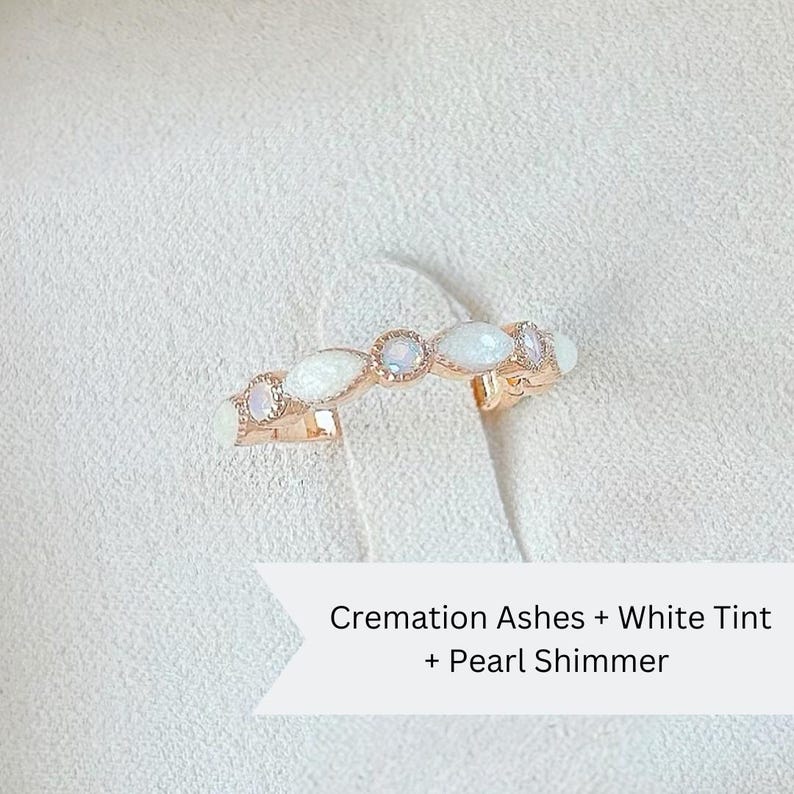 May include: A delicate rose gold ring with white opal stones set in a curved band. The ring is described as "Cremation Ashes + White Tint + Pearl Shimmer".
