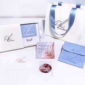 May include: A white box with a blue interior and the text "Pk jewellery" on the front. The box contains a small silver ring with a blue stone. The box is sitting on a white surface. There is a white bag with a blue ribbon and the text "Pk jewellery" on the front. There is a blue velvet pouch with the text "Pk jewellery" on the front. There is a white card with the text "Preserved memories worn close to the heart" on the front. There is a clear plastic bag with a white circle with the text "Breast milk" on the front.