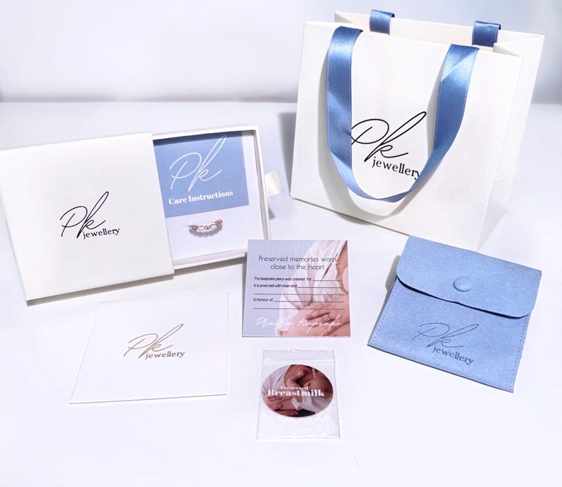 May include: A white box with a blue interior and the text "Pk jewellery" on the front. The box contains a small silver ring with a blue stone. The box is sitting on a white surface. There is a white bag with a blue ribbon and the text "Pk jewellery" on the front. There is a blue velvet pouch with the text "Pk jewellery" on the front. There is a white card with the text "Preserved memories worn close to the heart" on the front. There is a clear plastic bag with a white circle with the text "Breast milk" on the front.