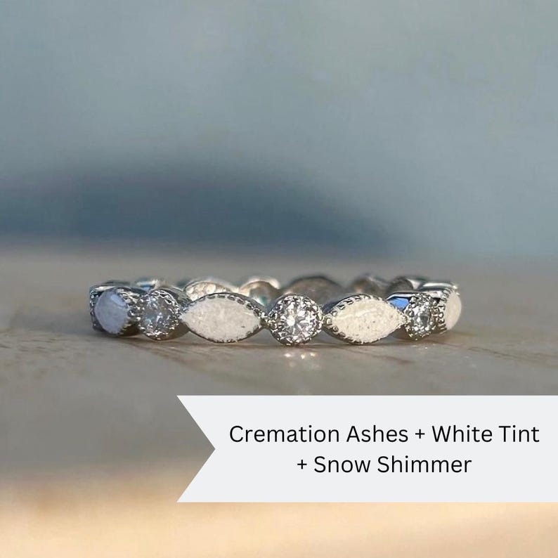 May include: A white gold band ring with small diamonds and white cremation ashes embedded in the band. The text on the image reads "Cremation Ashes + White Tint + Snow Shimmer".
